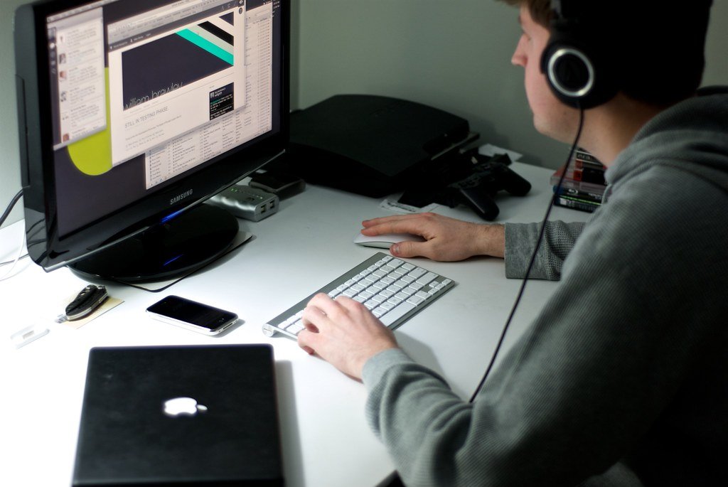 Persona con auriculares trabajando frente a un monitor, un teclado inalámbrico y una laptop cerrada sobre un escritorio blanco